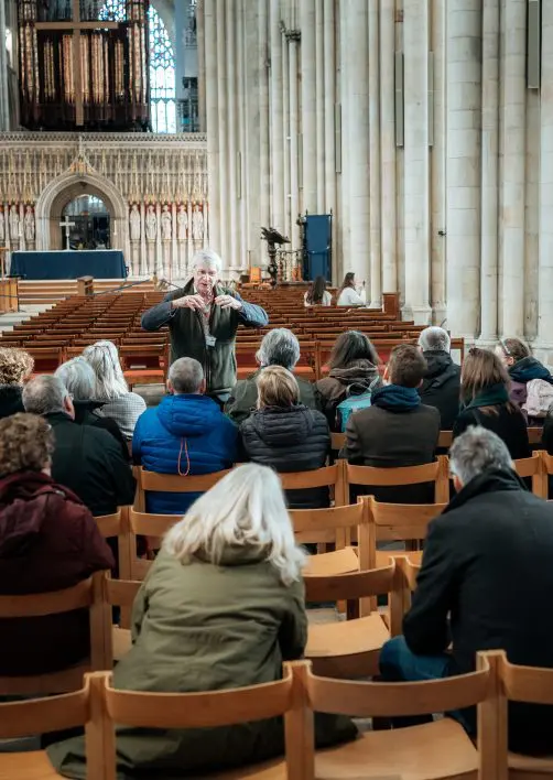 Inside York Minster cathedral, a Minster tour guide is presenting to a large group of people who are seated.