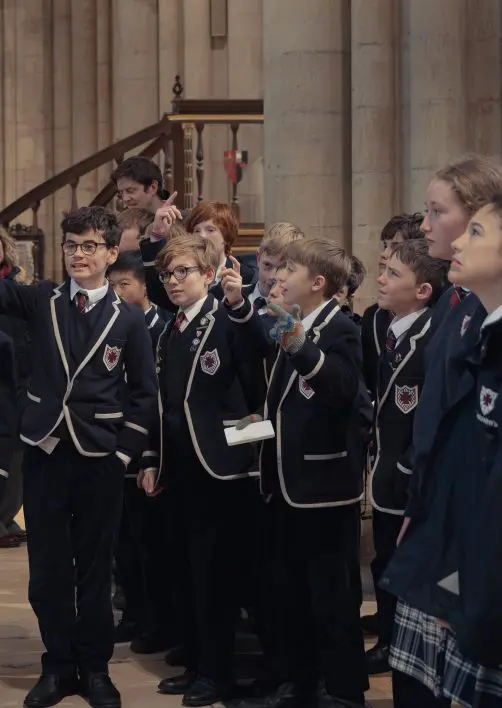 A group of school children on a school trip to York Minster, listening to the host. One child has their hand raised to answer a question.