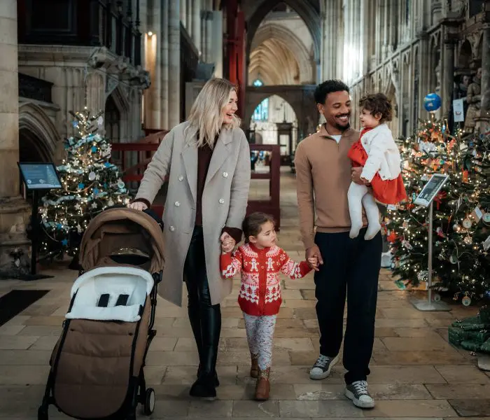 A family walk down the North Quire aisle in York Minster. One parent pushes a buggy, another carries a baby, both hold the hand of the elder child.